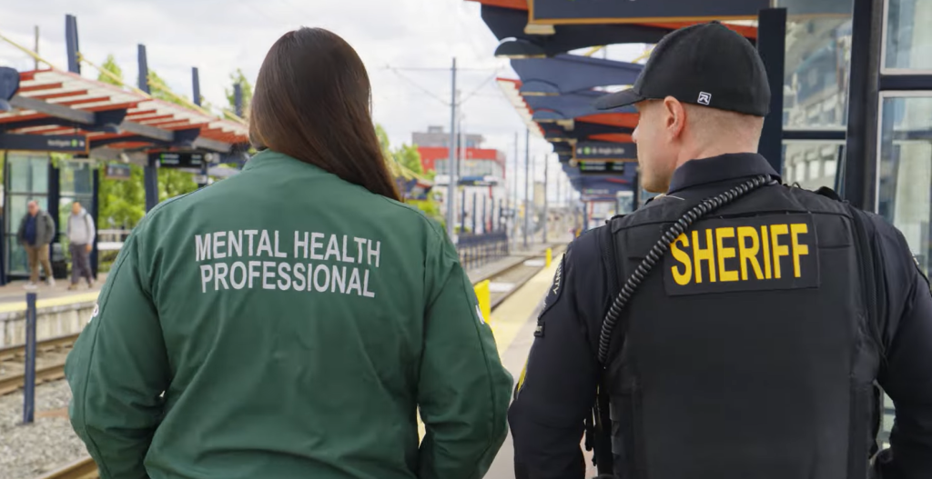 A woman wearing a green jacket with "mental health professional" on the back walks with a King County Sheriff's deputy at a light rail station.